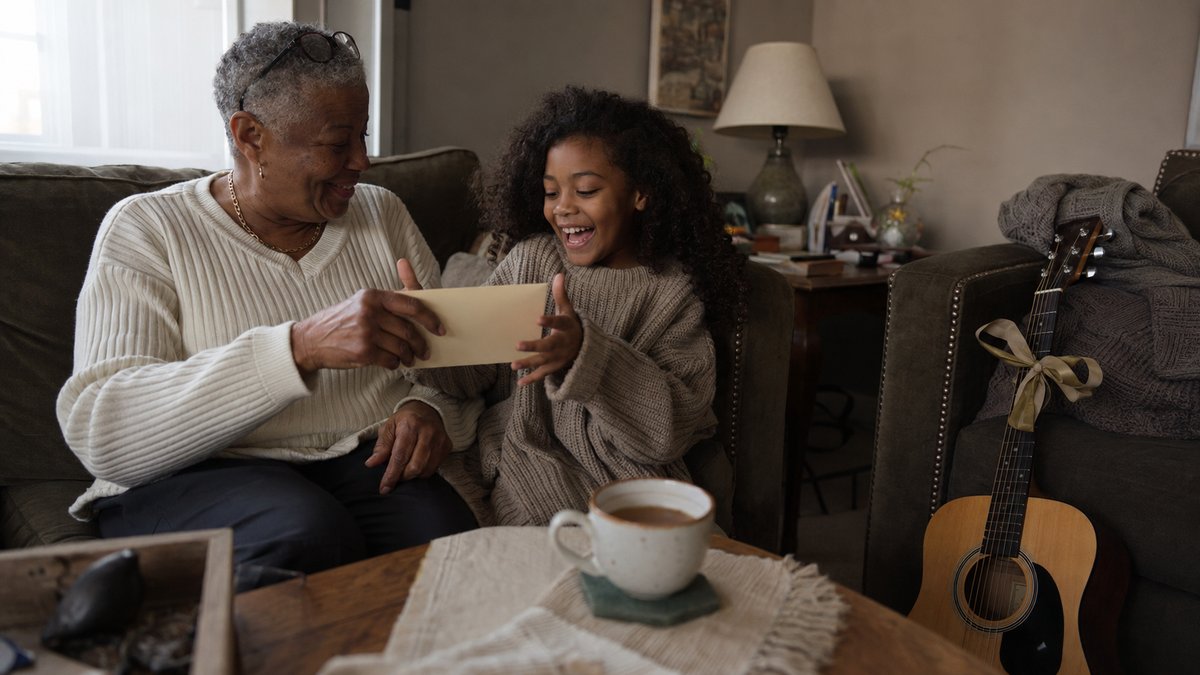 Grandmother handing her granddaughter a gift envelope on a sofa, with an acoustic guitar nearby