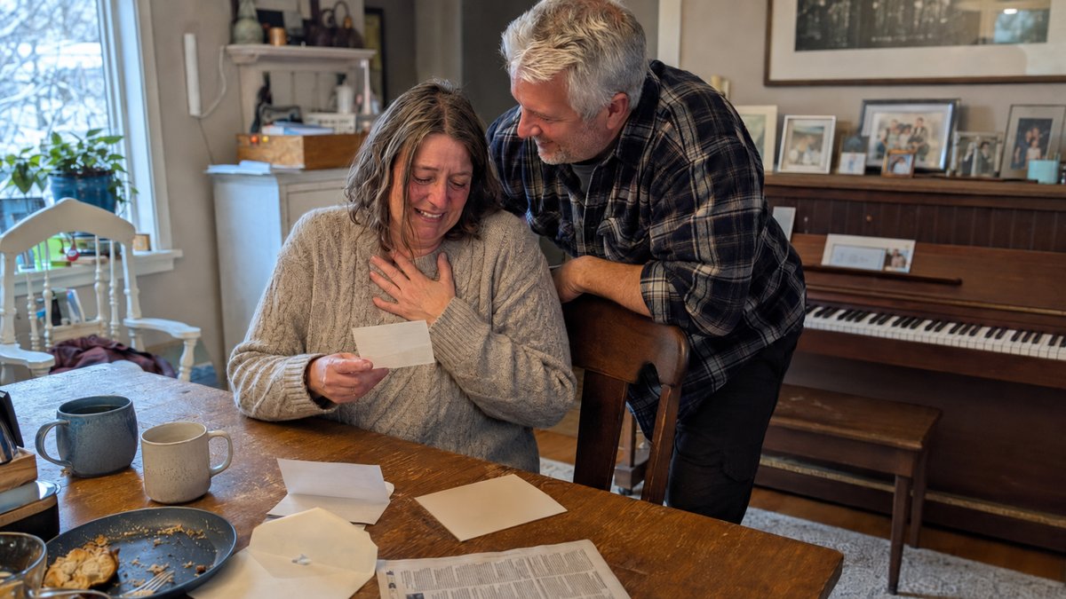 Woman in her fifties tearing up over a gift note at a kitchen table, upright piano behind
